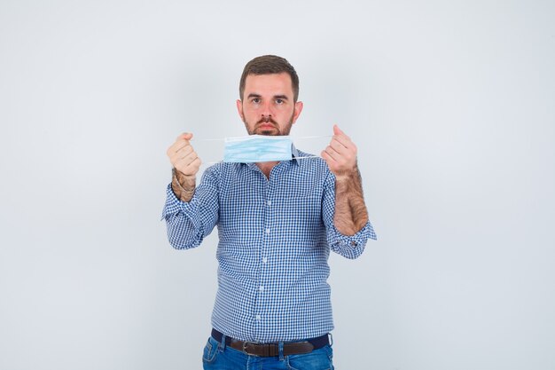 Handsome man holding mask in both hands in shirt, jeans and looking serious , front view.