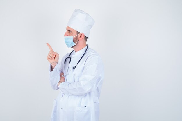 Handsome man holding stethoscope around neck, pointing left in white medical lab coat, mask and looking confident. front view.