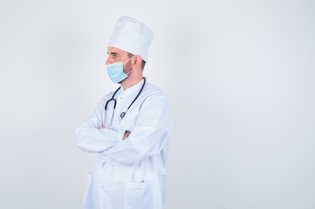 Handsome man holding stethoscope around neck, standing arms crossed in white medical lab coat, mask and looking confident. front view.