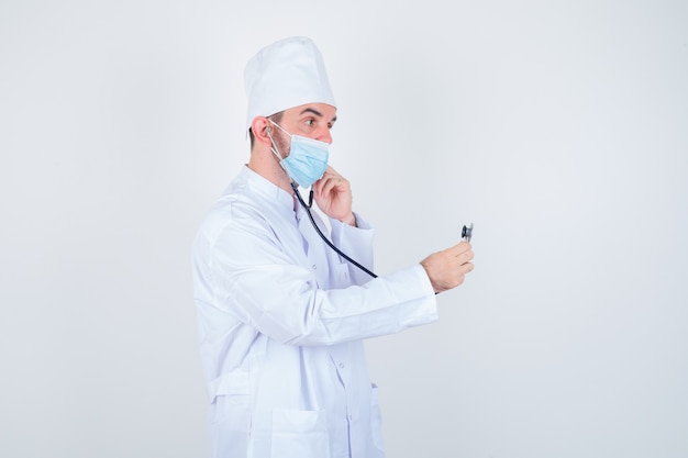 Красивый мужчина в белом халате медицинской лаборатории, маска, держащая в ушах стетоскоп, как будто он слушает и смотрит сосредоточенно, вид спереди.