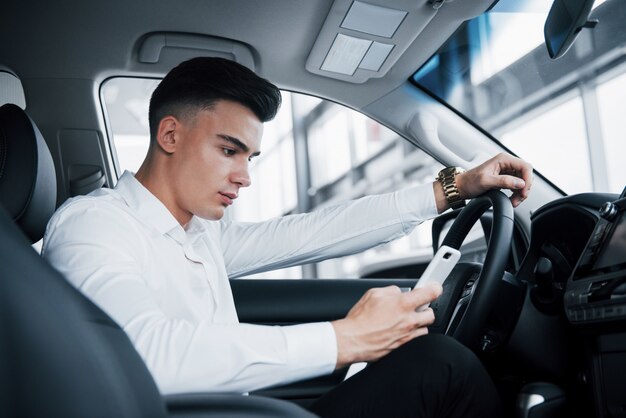 The handsome man is a buyer sitting in a car with a phone in a new car at a dealer center.