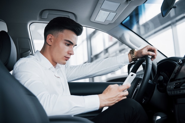 The handsome man is a buyer sitting in a car with a phone in a new car at a dealer center.