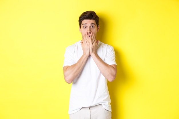 Handsome man looking shocked and speechless, holding hands on mouth, standing over yellow background