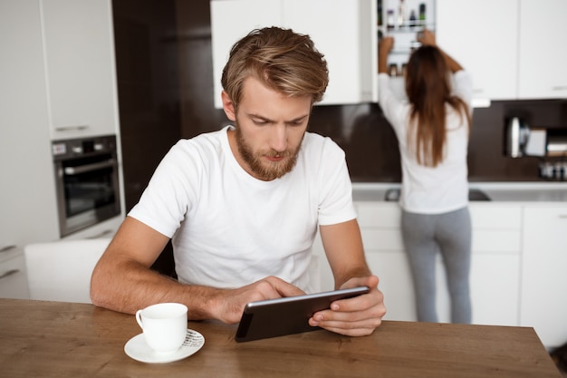 Handsome man looking at tablet. Girlfriend on the kitchen 