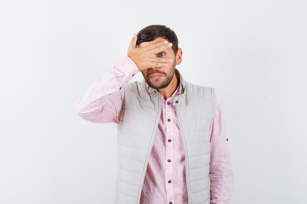 Handsome man looking through fingers in vest, shirt and looking scared.