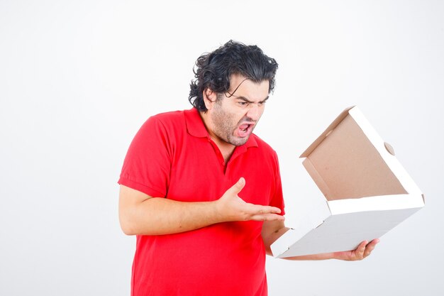 ハンサムな男が紙箱を開けて、赤いTシャツを着て怒った方法でそれに向かって手を伸ばし、怒っているように見える、正面図。