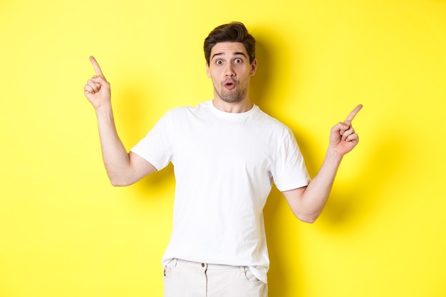 Handsome man pointing fingers sideways, showing two promos, standing over yellow background