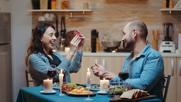 Handsome man proposing to his girlfriend marriage during festive dinner, in kitchen sitting at the table drinking a glass of red wine. Happy surprised woman smiling and hugging him.