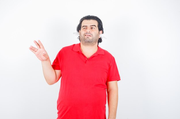 Handsome man raising hand, winking, standing with napkins in ears in red t-shirt and looking indecisive. front view.