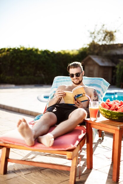 Handsome man reading book, lying on chaise near swimming pool