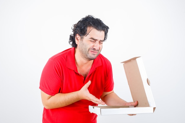 Handsome man in red t-shirt opening paper box, stretching hand toward it with disappointed manner and looking dismal , front view.