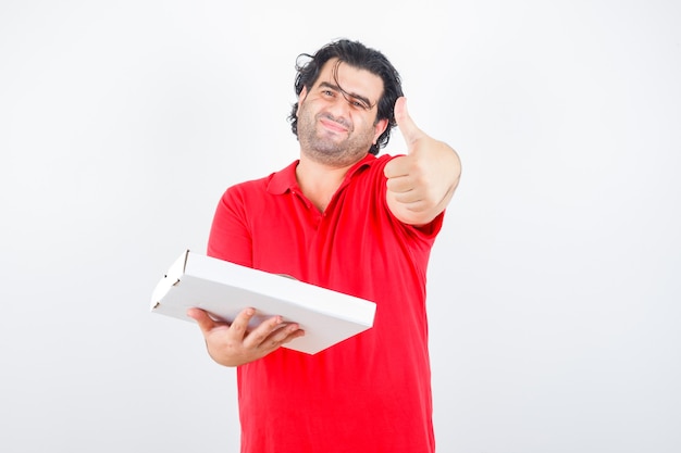 Красивый мужчина показывает палец вверх, держит бумажную коробку в красной футболке и выглядит веселым, вид спереди.