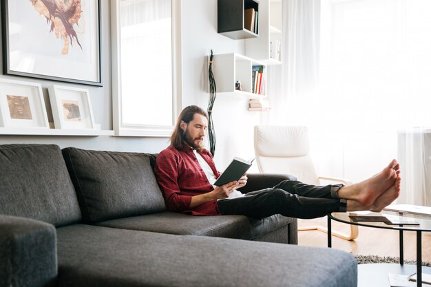 Красивый мужчина сидит и читает книгу на диване у себя дома
