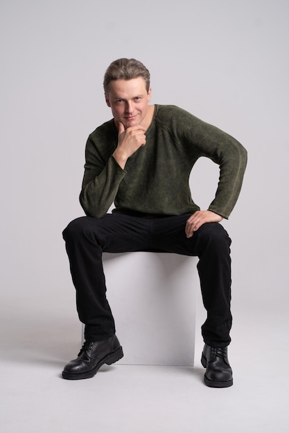 Handsome man sitting on the white cube with hand touching his chin looking at camera and smile wearing black denim and greengray knitted jumper on white background