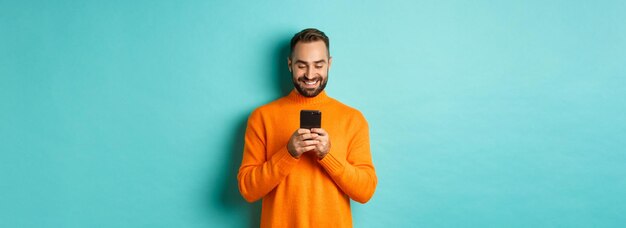 Handsome man smiling and texting message on mobile phone communicating online standing over light bl