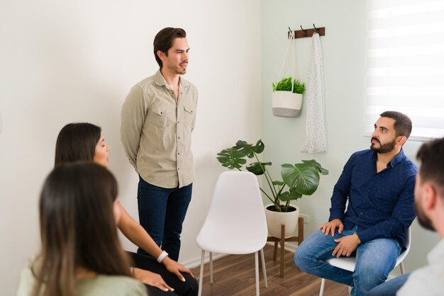 Handsome man standing in front of a circle of people and talking about his mental health issues. Young man in his 20s seeking group counseling