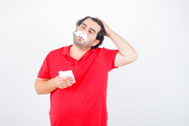 Handsome man standing with napkins in nostrils, holding napkin in hand, holding hand on head in red t-shirt and looking exhausted. front view.