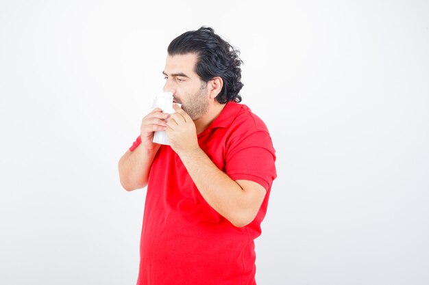 Handsome man standing with napkins in nostrils, holding napkin in hands in red t-shirt and looking exhausted. front view.