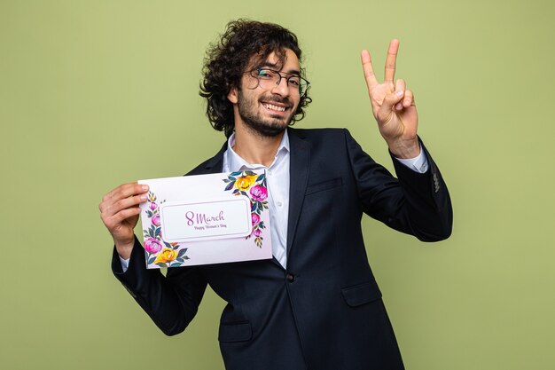 Handsome man in suit holding greeting card looking smiling cheerfully showing v-sign, celebrating international women's day march 8