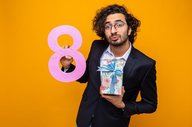 Handsome man in suit holding present and number eight looking happy and positive keeping lips like going to kiss celebrating international women's day march 8