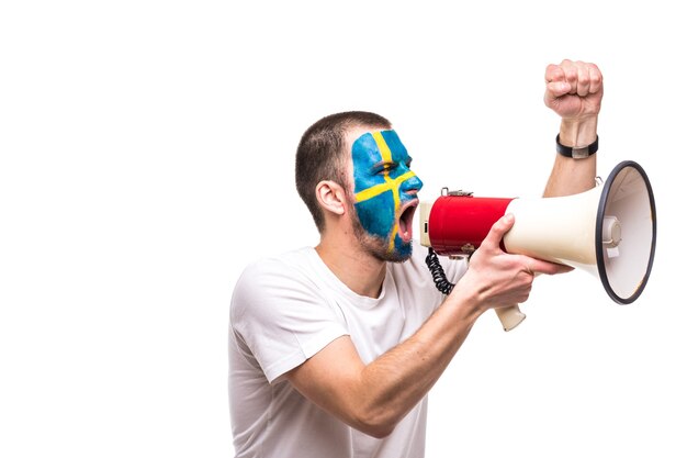 Handsome man supporter loyal fan of Sweden national team with painted flag face get happy victory screaming into megaphone with pointed hand. Fans emotions.