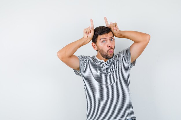 Handsome man in t-shirt keeping fingers as bull horns, pouting lips and looking humorous , front view.