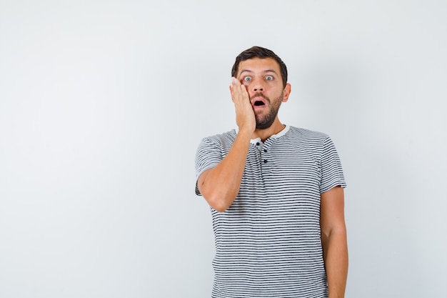 Handsome man in t-shirt keeping hand on cheek, opening mouth and looking terrified , front view.