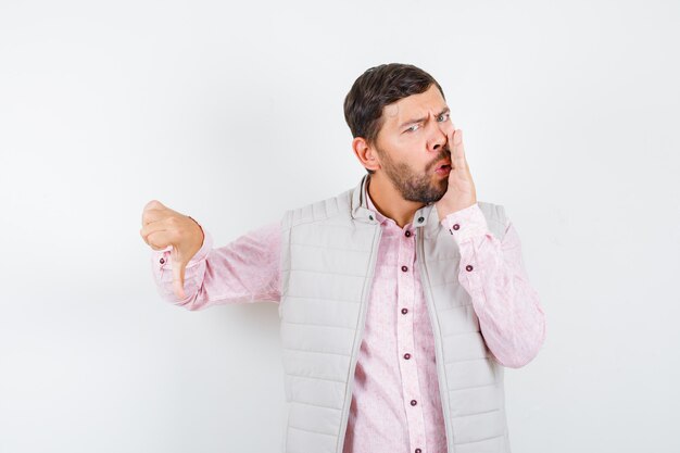 Handsome man thumb down, saying secret by keeping hand near mouth in vest, shirt and looking concerned .