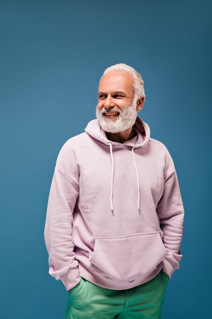 Handsome man in trendy hoodie posing on blue background Grey haired adult guy with beard in lilac sweatshirt and cool green pants smiling