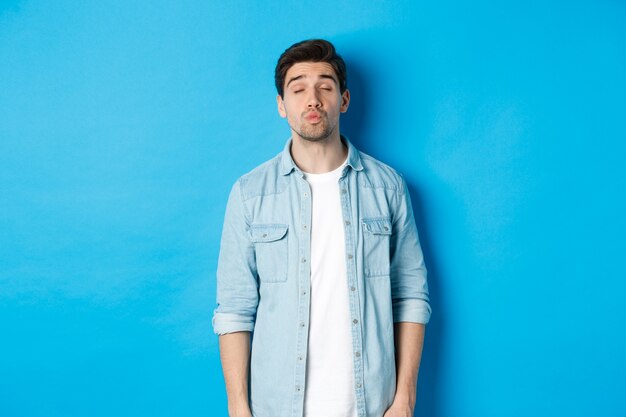 Handsome man waiting for kiss, pucker lips and close eyes while standing against blue background