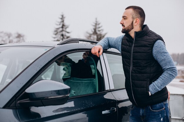 Handsome man in warm jacket standing by car covered with snow