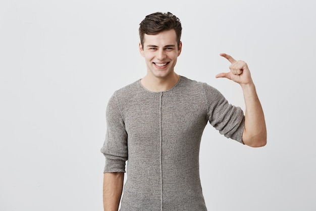 Handsome man wearing sweater showing something little with hands while gesturing and broadly smiling. Dark-haired caucasian male model demonstrating size of something 