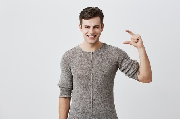 Handsome man wearing sweater showing something little with hands while gesturing and broadly smiling. Dark-haired caucasian male model demonstrating size of something 