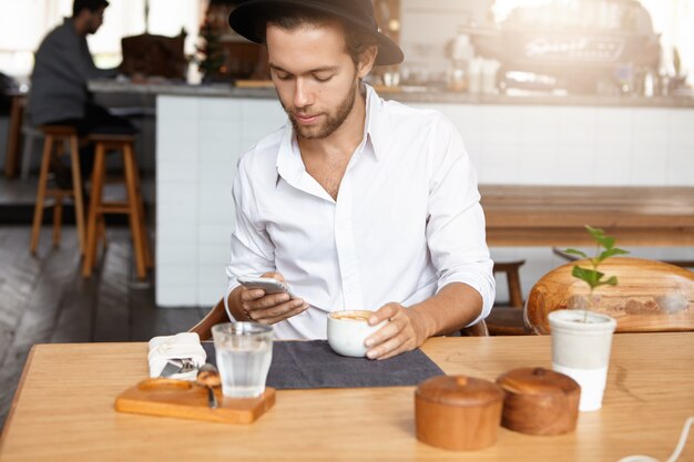 그의 휴대 전화에 무선 인터넷 연결을 사용하여 흰색 셔츠와 검은 세련된 모자를 쓰고 잘 생긴 남자, 아늑한 카페에서 테이블에 앉아있는 동안 소셜 네트워크를 통해 온라인으로 친구에게 메시지 보내기
