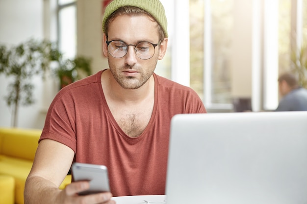 Handsome man wears trendy round spectacles, hat and t shirt, textes message on smart phone