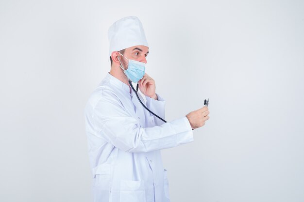 Handsome man in white medical lab coat, mask holding ear pieces of stethoscope in his ears as though he is listening and looking focused , front view.