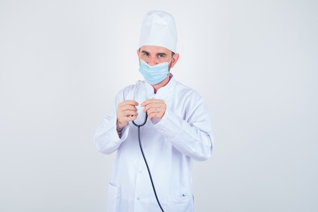 Handsome man in white medical lab coat, mask holding stethoscope and looking confident , front view.