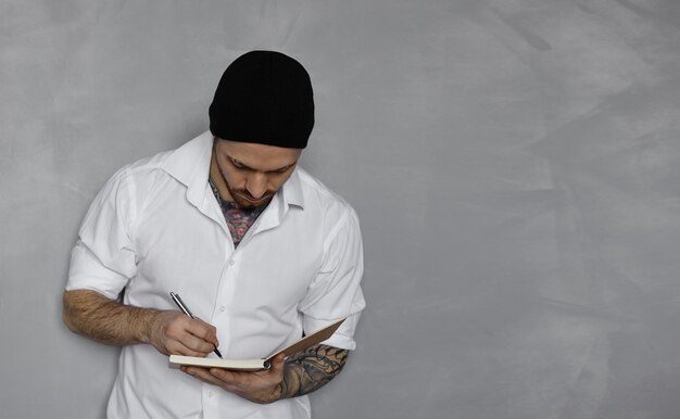 Handsome man in white shirt and black hat stay near grey wall and write notes in notepad