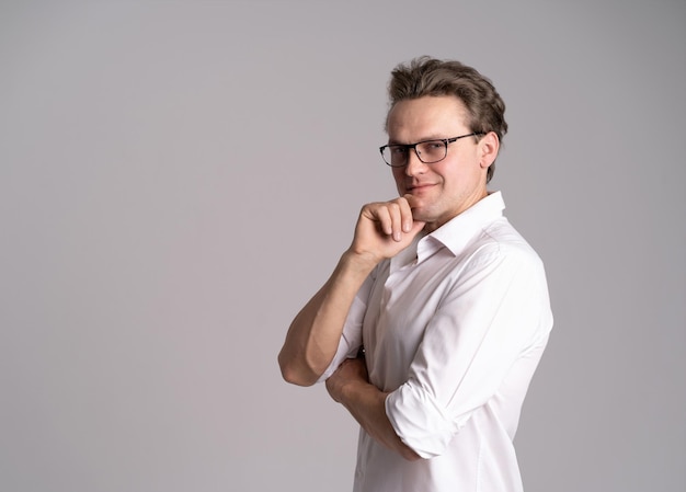 Handsome man in white shirt and eye glasses smiling standing sideways with a happy and confident positive expression with his arms crossed touching chin on a white background