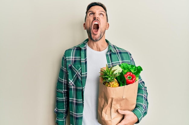 Free photo handsome man with beard holding paper bag with groceries angry and mad screaming frustrated and furious shouting with anger rage and aggressive concept