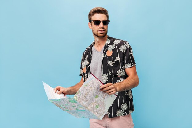 Handsome man with black sunglasses in cool summer shirt and light shirt looking away and holding map on isolated background