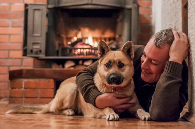 犬と家でカーペットに座っているハンサムな男。ペットの犬の家で暖炉の前でリラックスしている成熟した男