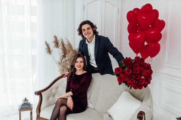 Handsome man with flowers standing behind his girlfriend sitting on the couch and looking at front