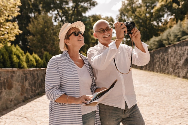 Handsome man with grey mustache in long sleeve shirt and jeans photographs and smiles with cool woman in hat glasses and blue striped blouse in park