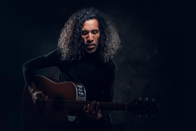 Handsome man with long hair is playing acoustic guitar at dark studio.