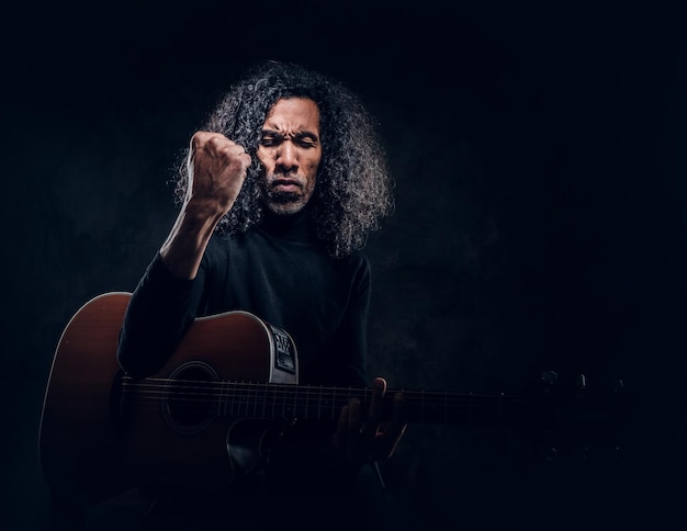 Handsome man with long hair is playing acoustic guitar at dark studio.