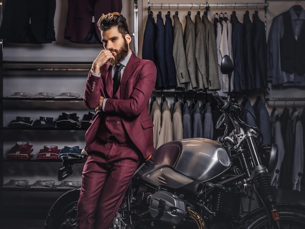 세련된 수염과 머리를 가진 잘생긴 남자는 빈티지 빨간 양복을 입고 남성 의류 매장의 복고풍 스포츠 오토바이 근처에서 포즈를 취합니다.