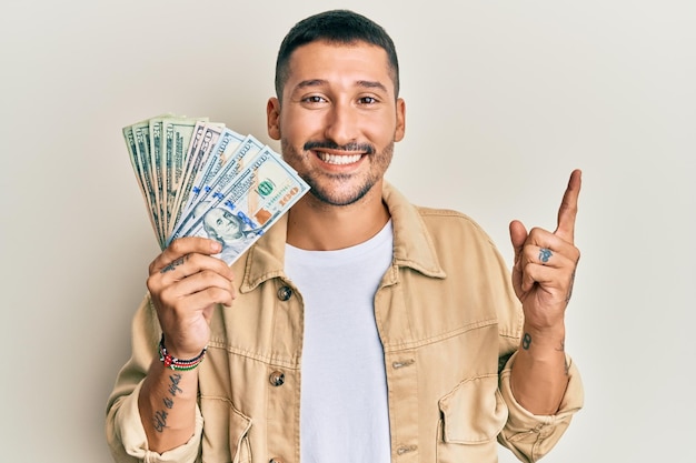 Handsome man with tattoos holding dollars smiling happy pointing with hand and finger to the side