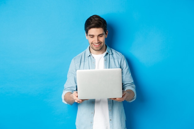 Handsome man working on laptop, smiling and looking at screen satisfied, standing against blue background