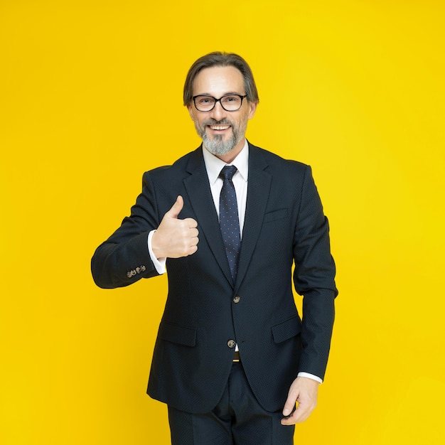 Красивый зрелый бизнесмен в черном костюме, показывающий большой палец вверх, деловой человек средних лет улыбается, глядя в камеру, изолированную на желтом фоне Бизнес-концепция